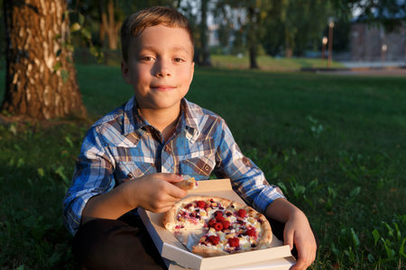 The boy eat a piece of pizza. Picnic on the grass. Boy have a dinner with sweet pizza on the meadow.