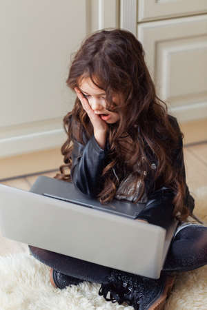 Small cute brunette caucasian baby sitting on floor on light artificial animal skin and sees something amazing on laptop.の写真素材