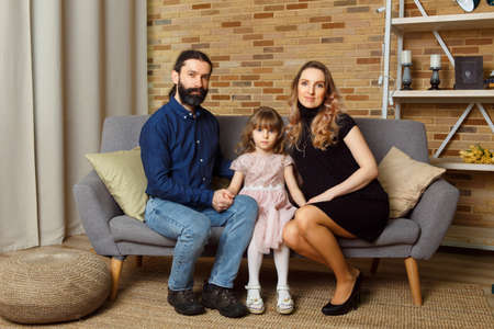 Happy young father, mother and daughter sit on wicker sofa at home. The image of a happy family expecting the second child, studioの写真素材