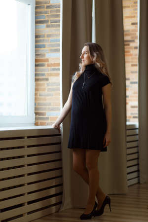 Thoughtful pregnant woman caressing her belly and looking out the window. Full length.の写真素材