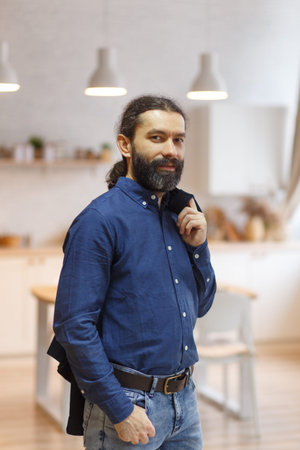 Portrait of a cute long-haired man with a beard in a blue shirt and jeans against the background of...