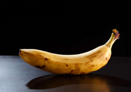 Single yellow banana on black background. Not perfect fruit with flaws and spots.の写真素材
