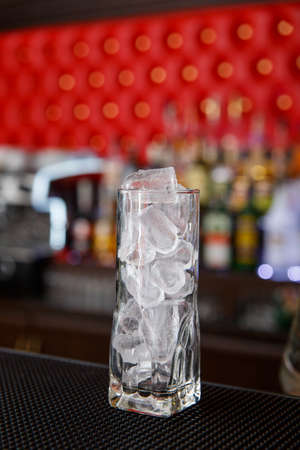A tall highball glass filled with ice cubes stands on the bar counter.の写真素材