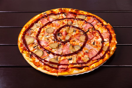 A whole rustic pizza with pickled cucumber, sousages, bacon and barbecue sauce is served on a paper plate on a wooden tableの写真素材