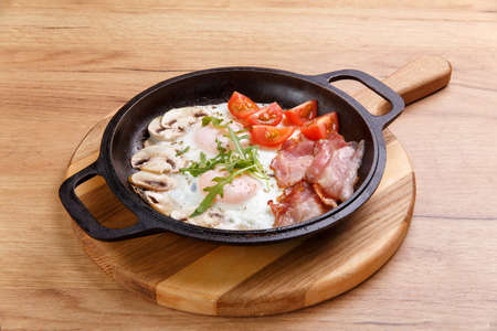 English breakfast in cooking pan with fried eggs, bacon, mushrooms and tomatoes on wooden standの写真素材