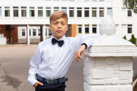 A beautiful eight-year-old boy in a school uniform, black trousers and a white shirt with bow tie poses in the school yard.の写真素材