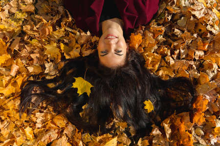 A happy young woman in red coat lies in the autumn leaves. Beautiful eyes, warm emotions, long black hair.の写真素材