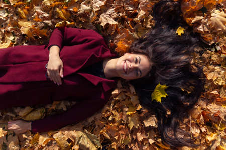 A happy young woman in red coat lies in the autumn leaves. Beautiful eyes, warm emotions, long black hair.の写真素材