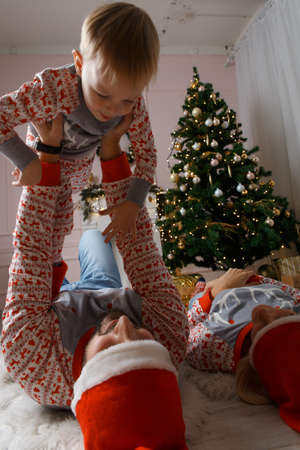 Happy family near christmas tree having fun on the floor. Christmas celebration at home. Mom, Dad and son weared in casual. Happy family funny playing on a bed. Christmas decorated grey interiorの写真素材