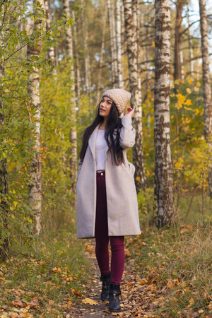 Portrait of a joyful young woman enjoying in the autumn park. Beautiful brunette girl in autumn grey coat and knitted hat. Relax in nature.の写真素材