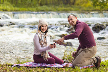 A beautiful young couple is resting on the banks of an autumn river and warm each other up with a hot drink.の写真素材