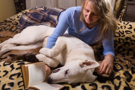 A woman lying on bed and reading a book with her companion white swiss shepherd dog. Love,...