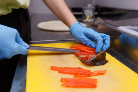 The hands of the cook in hygienic gloves with a sharp knife separate the salmon fillets from the bones. Cutting fresh fish in the kitchen on a cutting board.の写真素材