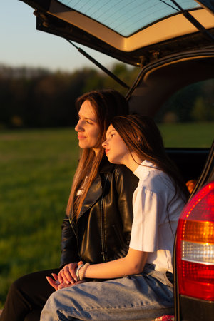 Mother and daughter camping on a hill and admiring the sunset while sitting in the car trunkの写真素材