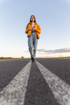 Cool modern teen girl poses on a lonely road while the sky background looks blue with clouds at sunset.の写真素材