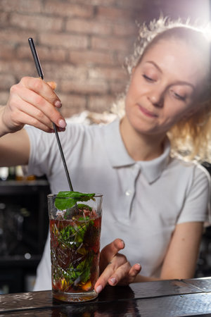 Young beautiful female bartender mixing a cocktail in a high glass. Mint petals are added to the cocktail.の写真素材