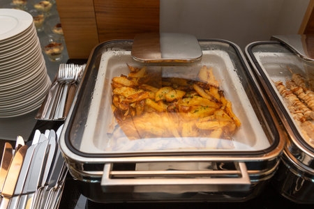 Fried potatoes garnish in a food warmer on a buffet table in a hotel restaurant. Table with dishware and shiny marmites waiting for guests.の写真素材