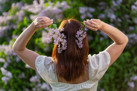 A girl with loose hair holds lilac flowers in her hands and stands with her back to the camera.の写真素材
