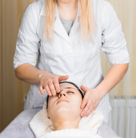 Beauty And Relaxation. Young woman enjoying facial massage, lying on spa bed indoor at beauty salon. Vertical shot of lady relaxing during facebuilding treatmentの写真素材