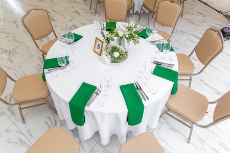 Wine glasses, silver tableware and green napkins on round tables covered with white tablecloth....