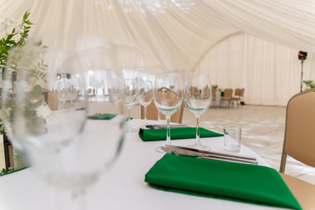 Close up of wine glasses, silver tableware and green napkins on round tables covered with white tablecloth. Catering concept. Interior of tent for wedding dinner, ready for guests.の写真素材