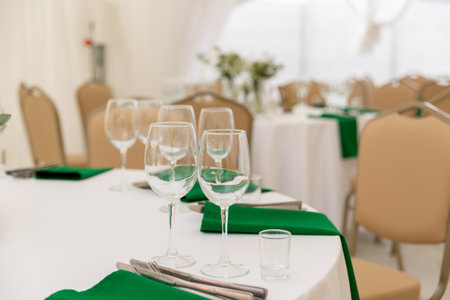 Close up of wine glasses, silver tableware and green napkins on round tables covered with white tablecloth. Catering concept. Interior of tent for wedding dinner, ready for guests.の写真素材