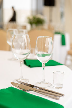 Close up of wine glasses, silver tableware and green napkins on round tables covered with white tablecloth. Catering concept. Interior of tent for wedding dinner, ready for guests.の写真素材
