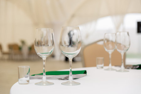 Close up of wine glasses, silver tableware and green napkins on round tables covered with white tablecloth. Catering concept. Interior of tent for wedding dinner, ready for guests.の写真素材
