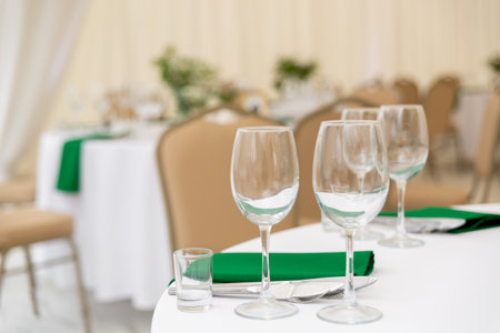 Close up of wine glasses, silver tableware and green napkins on round tables covered with white tablecloth. Catering concept. Interior of tent for wedding dinner, ready for guests.の写真素材