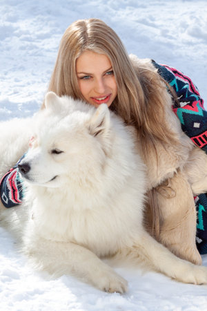 A young blonde with long hair poses in the park in the snow with a white husky dog. The girl is...