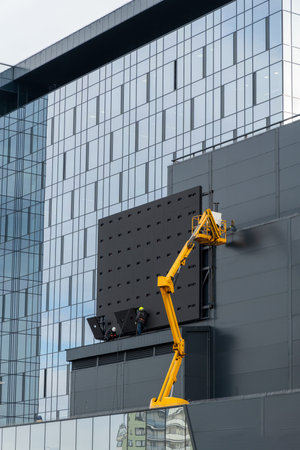 Two workers in hard hats mount elements of outdoor video advertising on a gray wall of a building...