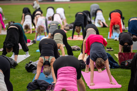Group yoga session on stadium lawn in shallow depth of field focusing on one practitioner. Connection, wellness, fitness community.の写真素材