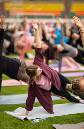 Group yoga session on stadium lawn in FYSM style, shallow depth of field focusing on one practitioner. Connection, wellness, fitness community.の写真素材