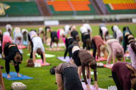 Group yoga session on stadium lawn in shallow depth of field focusing on one practitioner. Connection, wellness, fitness community.の写真素材