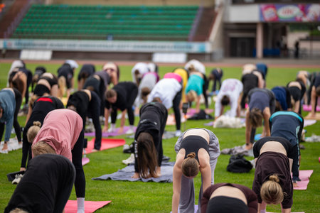 Group yoga session on stadium lawn in FYSM style, shallow depth of field focusing on one practitioner. Connection, wellness, fitness community.の写真素材