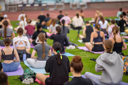 Group yoga session on stadium lawn in FYSM style, shallow depth of field focusing on one practitioner. Connection, wellness, fitness community.の写真素材