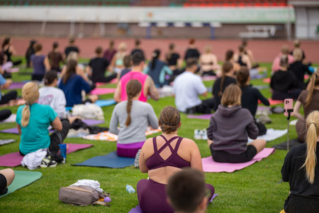 Group yoga session on stadium lawn in FYSM style, shallow depth of field focusing on one practitioner. Connection, wellness, fitness community.の写真素材
