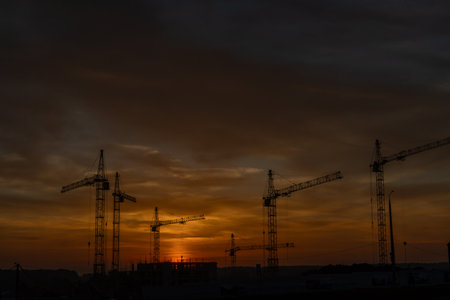Silhouettes of construction cranes on a building site against a sky lit by the setting sun.の写真素材