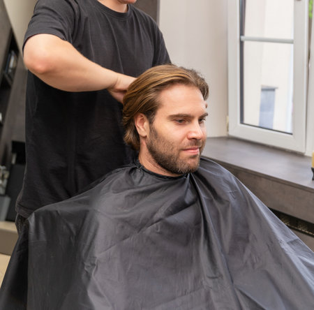 A hairdresser fastens a protective black cape on a male client before a haircut in a modern and clean salon. The client looks ahead, ready for the service to begin.の写真素材