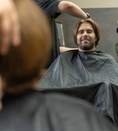 A hairdresser cuts a male client's long brown hair in a barbershop. The man sits with a neutral expression, wearing a black cape. A reflection is visible on the left.の写真素材