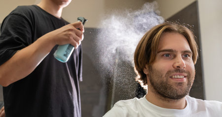 A barber sprays water on a male client's hair before a haircut. The client, with a smile, looks...