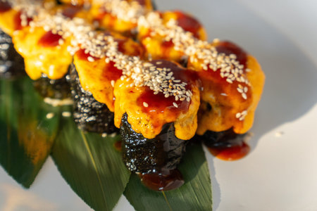 Close-up of baked sushi rolls topped with melted cheese, sauce, and sesame seeds, served on a green...