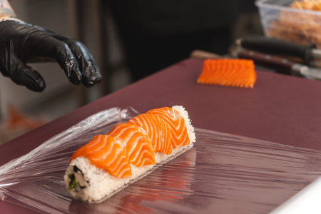 Chef in black glove preparing fresh salmon sushi roll on plastic wrap over cutting board in...