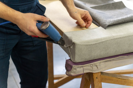 Worker's hand operates a blue pneumatic stapler. Grey patterned fabric is secured on surface....