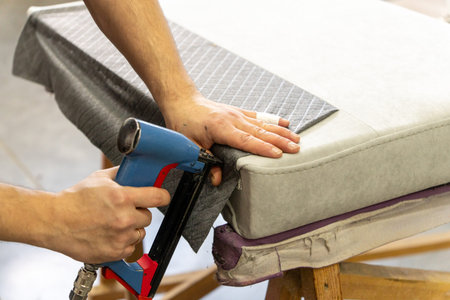 A worker uses a pneumatic stapler to attach grey fabric during furniture upholstery. Close-up of hands and tool in a workshop. Manual labor, repair, and craft concept.の写真素材