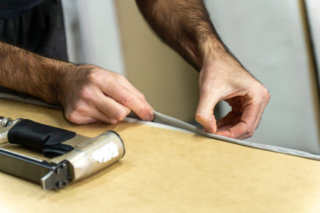 Close-up of upholsterer hands folding fabric edge on a wooden furniture frame, with staple gun nearby. Professional upholstery repair and DIY restoration work in a workshop.の写真素材