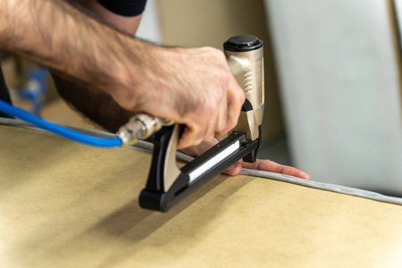 Close-up of upholsterer using a pneumatic staple gun to attach fabric to a wooden furniture frame. Professional workshop repair and DIY renovation concept with industrial tool.の写真素材