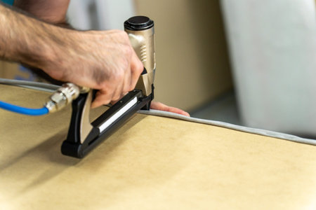 Close-up of upholsterer using a pneumatic staple gun to attach fabric to a wooden furniture frame. Professional workshop repair and DIY renovation concept with industrial tool.の写真素材