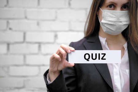 A European girl wearing a medical mask holds a sign with the inscription, against the backdrop of a white brick wall QUIZ - textの写真素材