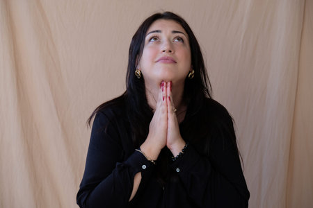 A woman with long dark hair sits, looks up and prays with folded hands to the higher powers